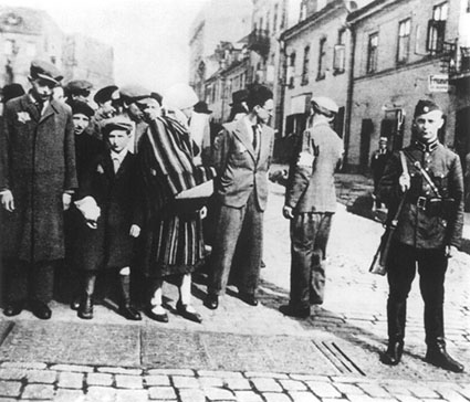 Saksalaissotilas poseeraa Varsovassa Juutalaispoliisimiehen (kuvassa selin käsivarsinauhan kanssa) käskyttäessä juutalaistovereita, jotka odottavat pääsyä Varsovan juutalaisosaan.
